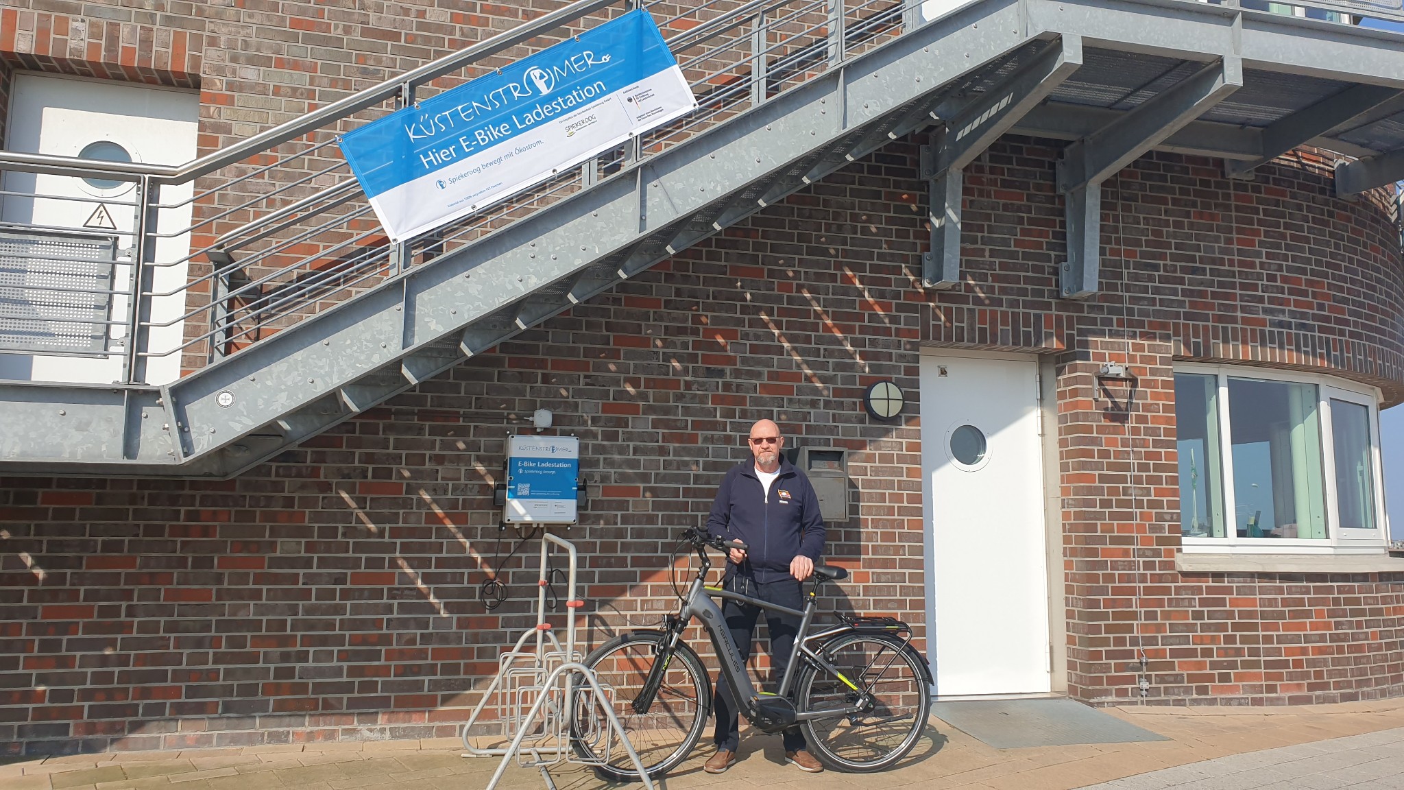 E-Bike Ladestation im Hafen Neuharlingersiel mit einem Mitarbeiter der Nordseebad Spiekerooog GmbH mit Fahrrad davor. 